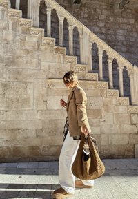 Femme marchant devant un escalier en pierre orné, portant un blazer beige, un pantalon blanc, des lunettes de soleil et tenant un grand sac en osier marron.