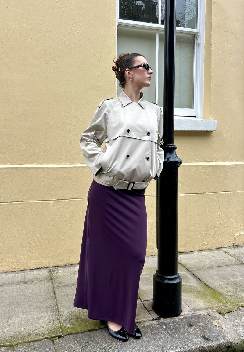 Frau trägt beige Trenchcoat und lila Rock, steht auf dem Bürgersteig neben einem schwarzen Laternenpfahl, mit den Händen in den Taschen, blickt nach rechts.