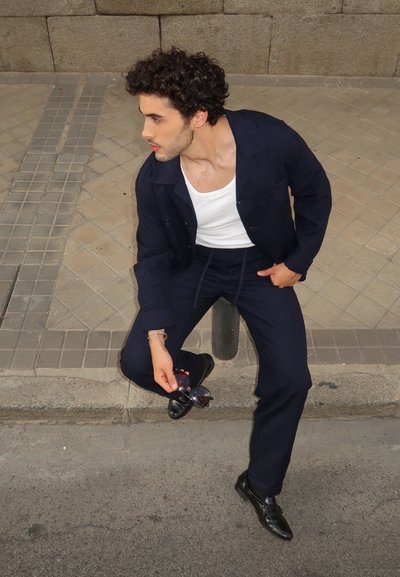 Chaqueta y pantalones azul marino, camiseta sin mangas blanca, zapatos negros. Sosteniendo gafas de sol negras, sentado en el pavimento cerca de un muro de piedra.