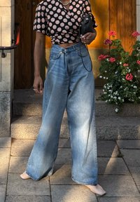 Personne portant un haut court à pois et un jean large bleu clair, tenant un sac noir, debout sur des marches en pierre près de fleurs.