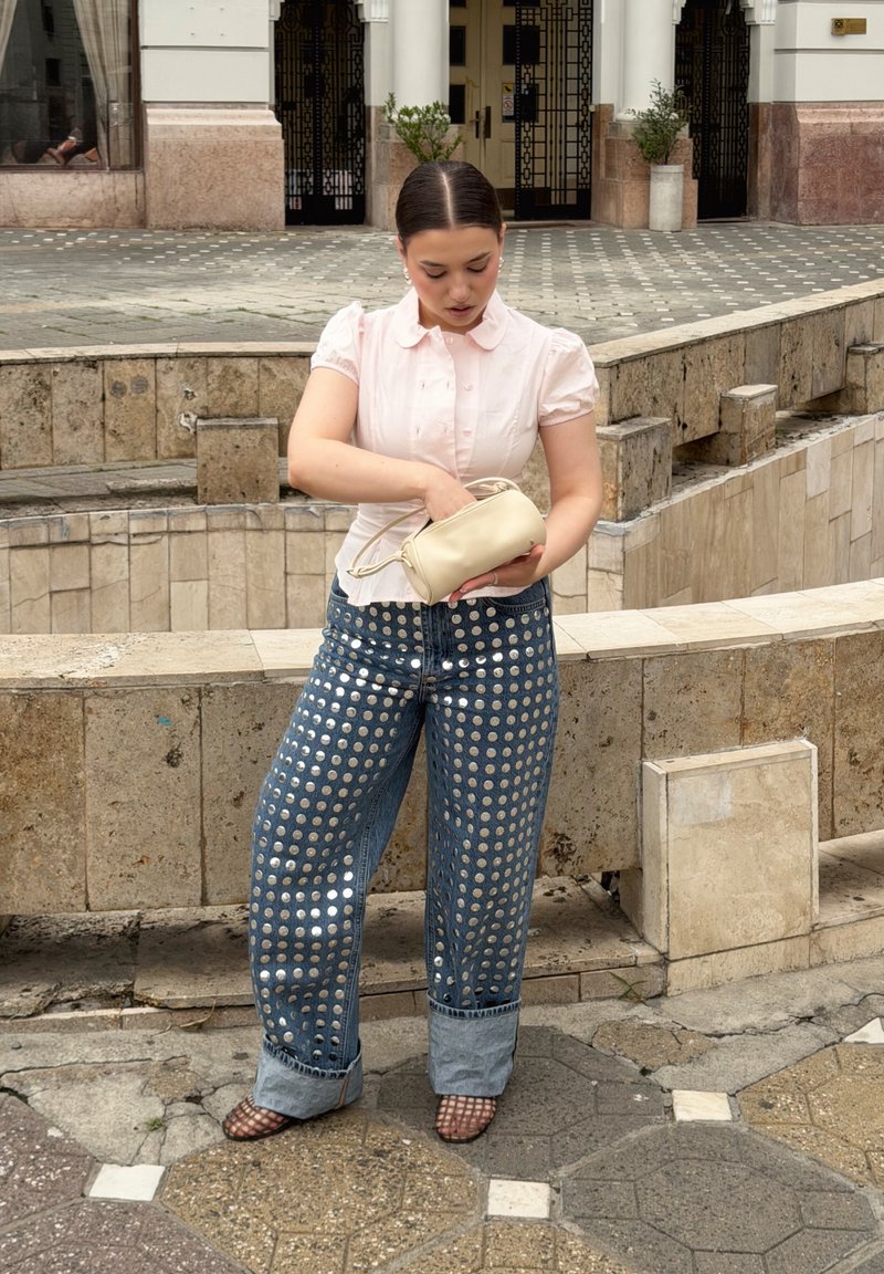 Jeune femme en blouse rose et jean à motifs de points réfléchissants se tient dehors, cherchant à l'intérieur d'un petit sac beige.