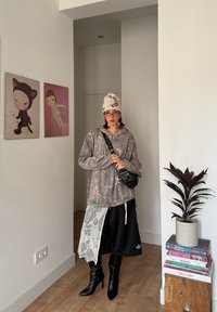Sweat à capuche zippé gris surdimensionné à motif, pantalon court noir avec dentelle, bottes noires à talons montant jusqu'au genou, et foulard blanc à motifs.