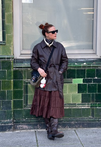 Abrigo de cuero marrón con cinturón sobre un suéter crema, combinado con una falda marrón oscuro con volantes y botas marrones hasta la rodilla. Bolso negro con charm.