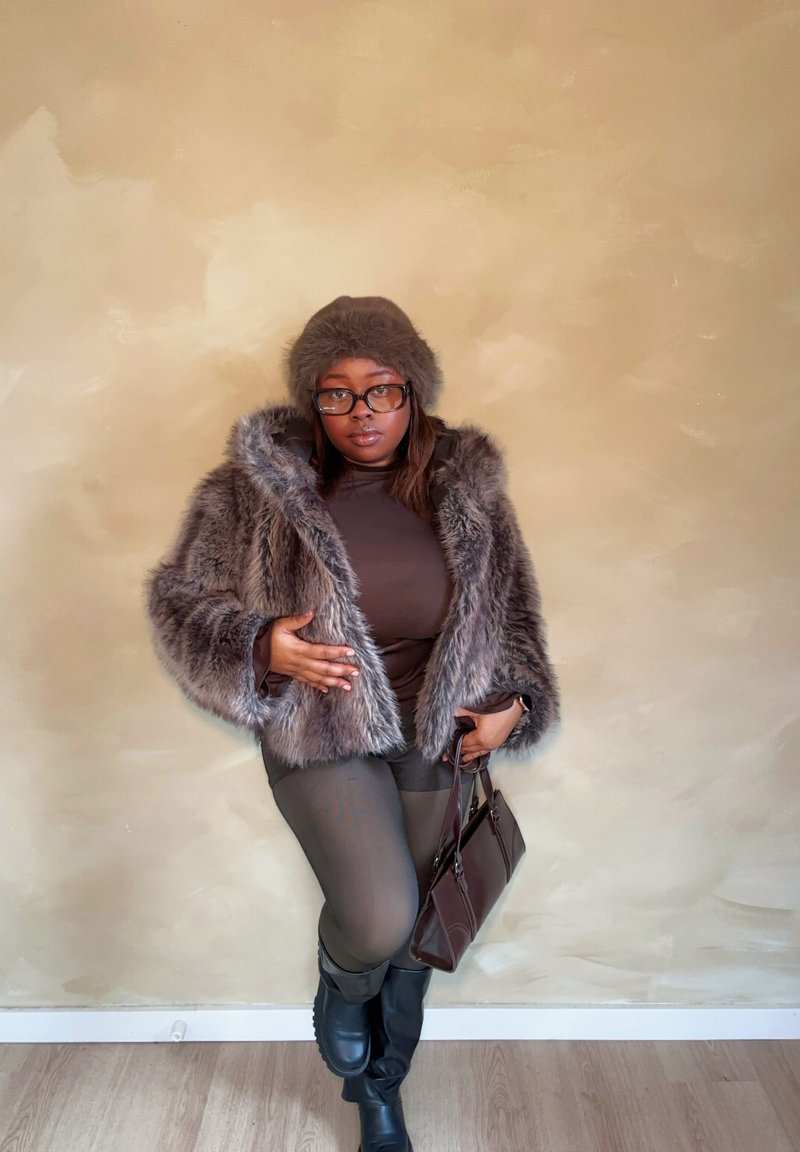 Manteau en fausse fourrure gris avec large col, assorti à un haut marron, des leggings gris et des bottes noires montant jusqu'aux genoux. Tenant un sac à main marron.