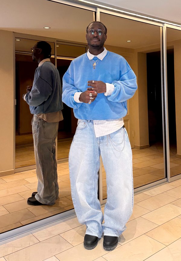 Homme portant un pull oversize bleu clair, une chemise blanche, un jean ample, des chaussures noires et des bijoux, debout à l'intérieur près d'un mur miroir.
