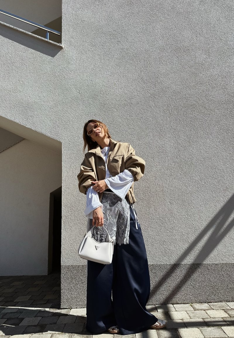 Femme portant une veste beige oversize, un chemisier blanc avec un détail en dentelle, un pantalon large bleu marine, des lunettes de soleil, tenant un sac à main blanc, debout contre un mur texturé.