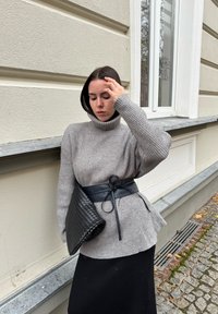 Grey ribbed turtleneck jumper with a black waist belt, holding a textured black clutch bag. Set against a light-coloured wall.