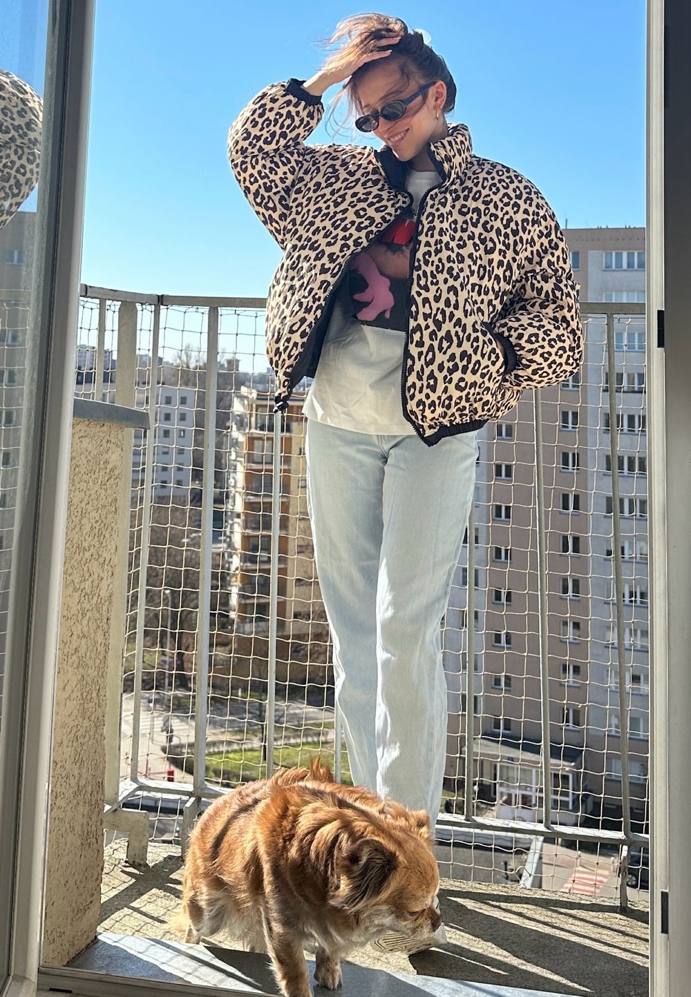 Luipaardprint pufferjack met een hoge kraag en rits, gecombineerd met lichtblauwe jeans. Een kleine bruine hond staat op het balkon. Heldere blauwe lucht.