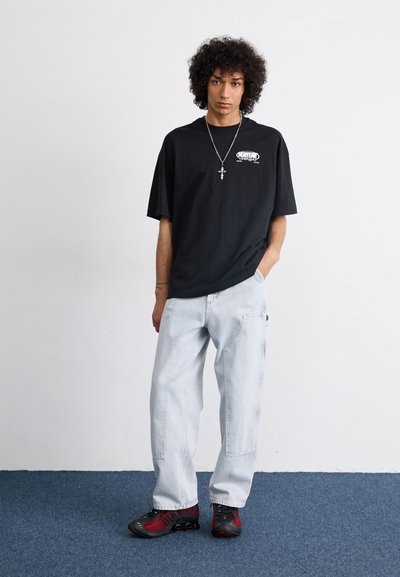 Camiseta de algodón negra de gran tamaño con estampado gráfico, vaqueros holgados azul claro y zapatillas negras/rojas. Collar de cruz plateado sencillo.