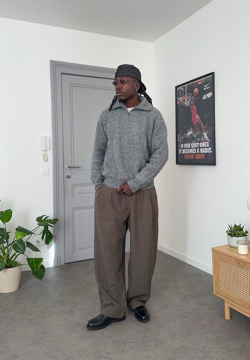 Un homme portant un pull gris, un pantalon brun et une casquette noire se tient à l'intérieur près d'une porte et d'une affiche de motivation sur le basketball accrochée au mur.