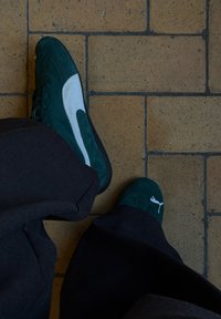 Grønne semskede sneakers med hvide detaljer og snørebånd, parret med mørke bukser, på et flisegulv.