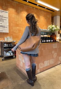 Tummaa nahkaa oleva olkalaukku, pehmeällä muodolla ja vähäisellä metalliosilla, harmaan bleiserin kanssa käytettynä, yhdistetty laskostettuun hameeseen ja polven yläpuolelle ulottuviin ruskeisiin saappaisiin.