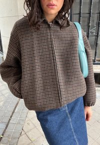 Femme portant une veste zippée à motif pied-de-poule marron, une jupe en jean bleue, et portant un sac à bandoulière bleu clair, debout sur un trottoir carrelé.