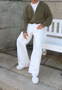 Sudadera con capucha verde oliva acanalada con cremallera, pantalones anchos blancos con rayas laterales y zapatillas blancas con detalles en negro. Anillos plateados en las manos.