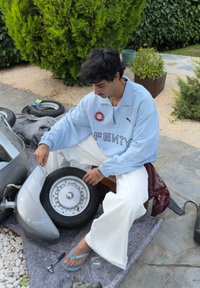 Scooter prateado com um pneu cinza, camisa de tecido azul e calças brancas. Ferramentas, incluindo uma chave inglesa e uma chave de catraca, estão visíveis em uma manta texturizada.