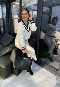 Λευκό ριπ πουλόβερ με ναυτικές ρίγες, κρέμα παντελόνια και μαύρη δερμάτινη τσάντα. Τοποθετημένο σε έναν μοντέρνο χώρο με μεγάλα παράθυρα και μαλακά μαξιλάρια.