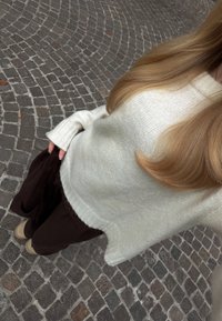 Personne aux longs cheveux blonds portant un pull en tricot blanc, un pantalon foncé et des chaussures beiges, debout sur un pavement en pavés.