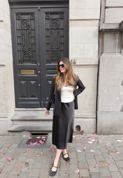 Blazer negro sobre una blusa blanca, combinado con una larga falda de satén negra. El conjunto incluye zapatos planos negros y un bolso pequeño. Pétalos rosas esparcidos sobre los adoquines.