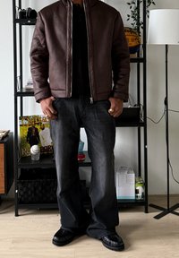 Veste aviateur en cuir marron avec fermeture éclair devant, polo noir, jean foncé ample et chaussures noires à enfiler avec détails texturés.