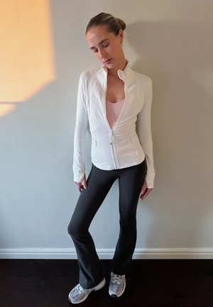 Chaqueta blanca con cremallera sobre una blusa rosa, combinada con pantalones negros acampanados y zapatillas deportivas grises. Tejido suave con un diseño ajustado y mangas largas.