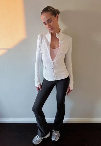 Chaqueta blanca con cremallera sobre una blusa rosa, combinada con pantalones negros acampanados y zapatillas deportivas grises. Tejido suave con un diseño ajustado y mangas largas.