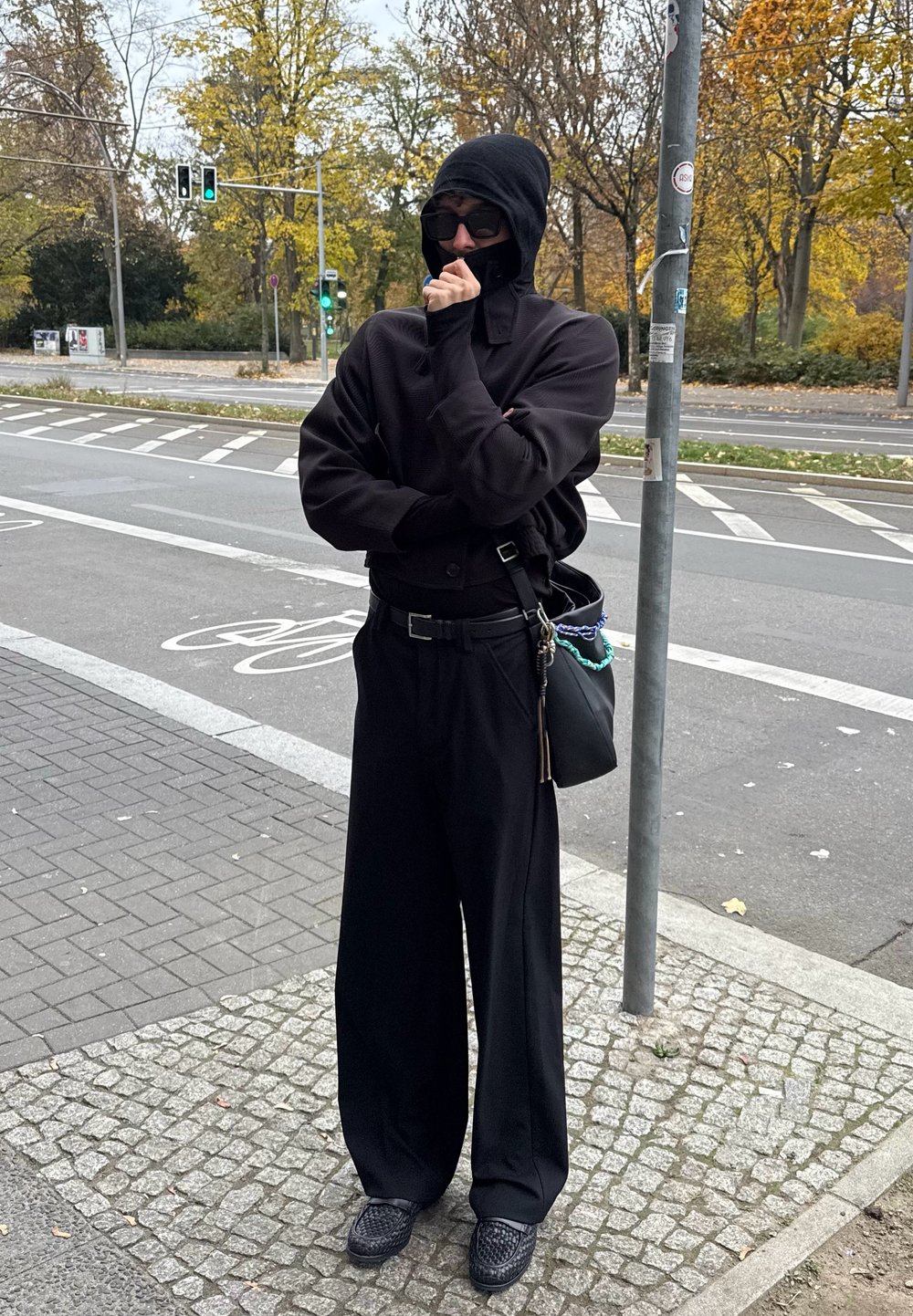 Μαύρος oversized πουλόβερ με ψηλό λαιμό, μαύρο παντελόνι με φαρδιά πόδια και υφασμάτινα μαύρα παπούτσια. Κρατά μια μαύρη τσάντα με αλυσίδες.