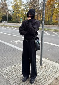 Μαύρος oversized πουλόβερ με ψηλό λαιμό, μαύρο παντελόνι με φαρδιά πόδια και υφασμάτινα μαύρα παπούτσια. Κρατά μια μαύρη τσάντα με αλυσίδες.