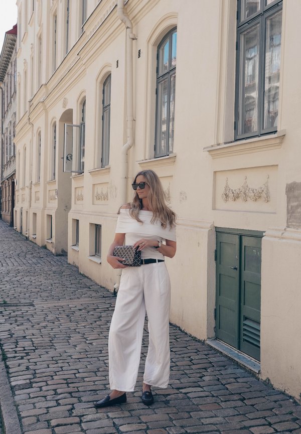 Mujer con atuendo blanco y gafas de sol está de pie en una calle empedrada junto a un edificio beige con ventanas altas y puerta verde.