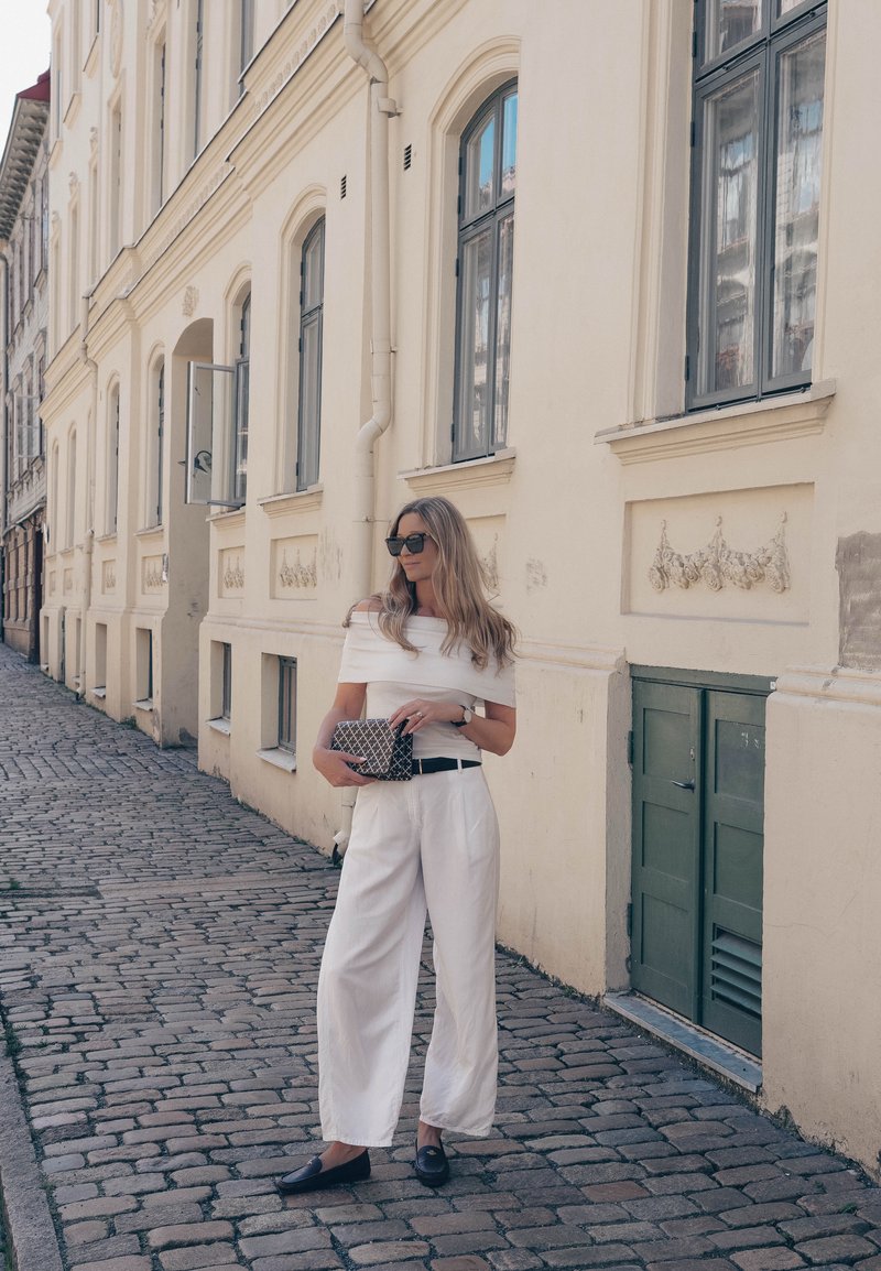 Femme portant une tenue blanche et des lunettes de soleil, debout dans une rue pavée à côté d'un bâtiment beige avec de grandes fenêtres et une porte verte.