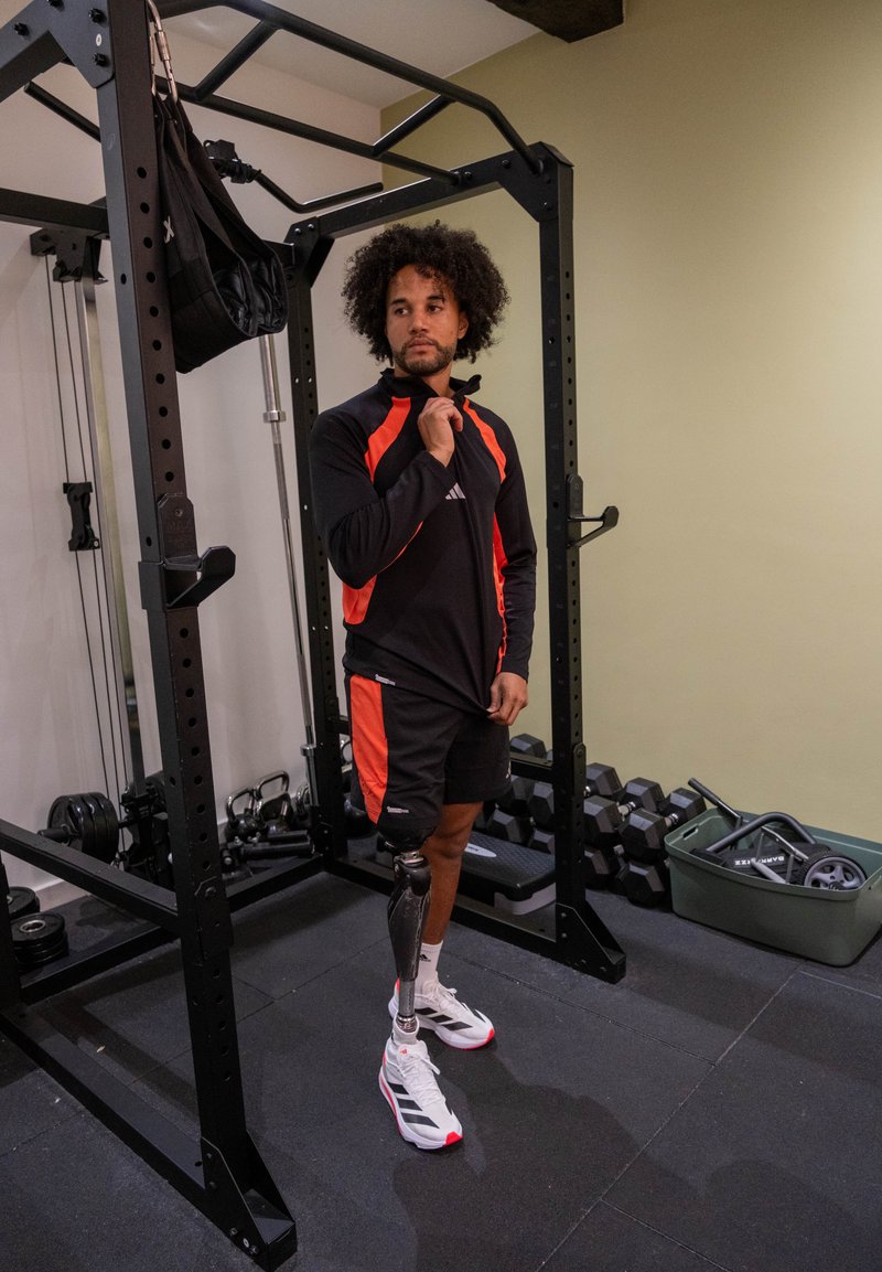 Homem com perna protética vestido com roupa desportiva preta e vermelha, em pé no ginásio perto de um suporte para levantamento de pesos e halteres.