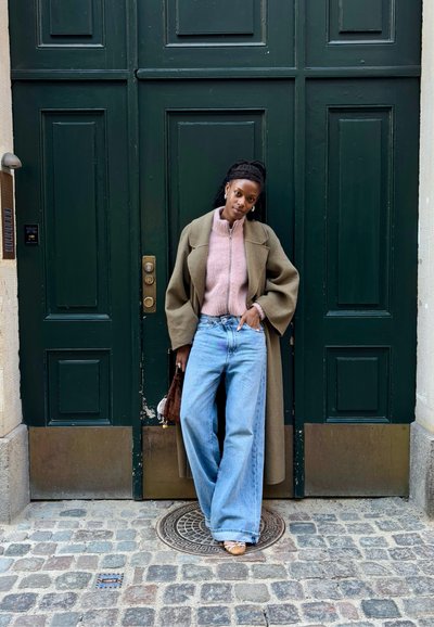 Abrigo oversize color verde oliva sobre un suéter rosa claro con cremallera, combinado con jeans de pierna ancha azules y un bolso marrón. Fondo de puerta verde.