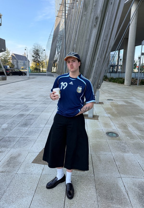 Joven con camiseta deportiva azul y pantalones anchos negros sosteniendo una taza de café, de pie en una plaza embaldosada con estructuras arquitectónicas modernas.