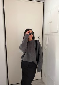 Pull gris texturé, jupe noire et lunettes de soleil noires. Debout contre une porte de couleur claire avec des éléments environnants minimalistes.