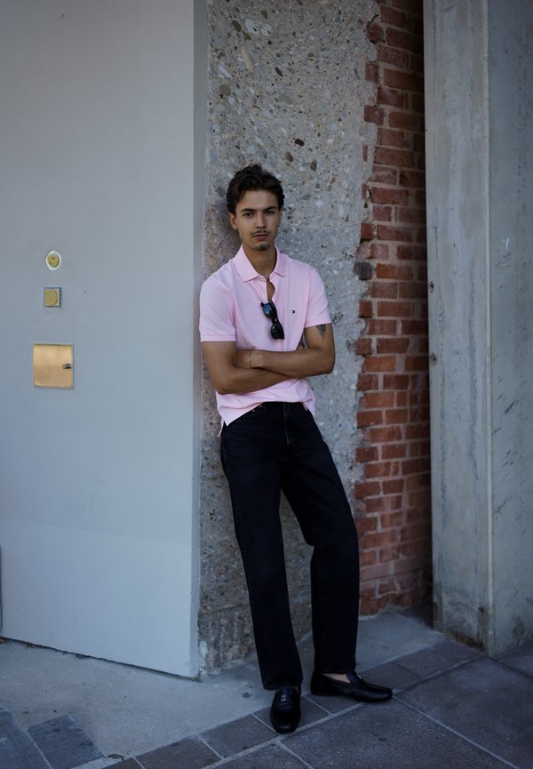 Hombre con camiseta polo rosa y pantalones negros se apoya contra una pared de piedra y ladrillo mezclados con los brazos cruzados y gafas de sol colgando del cuello.