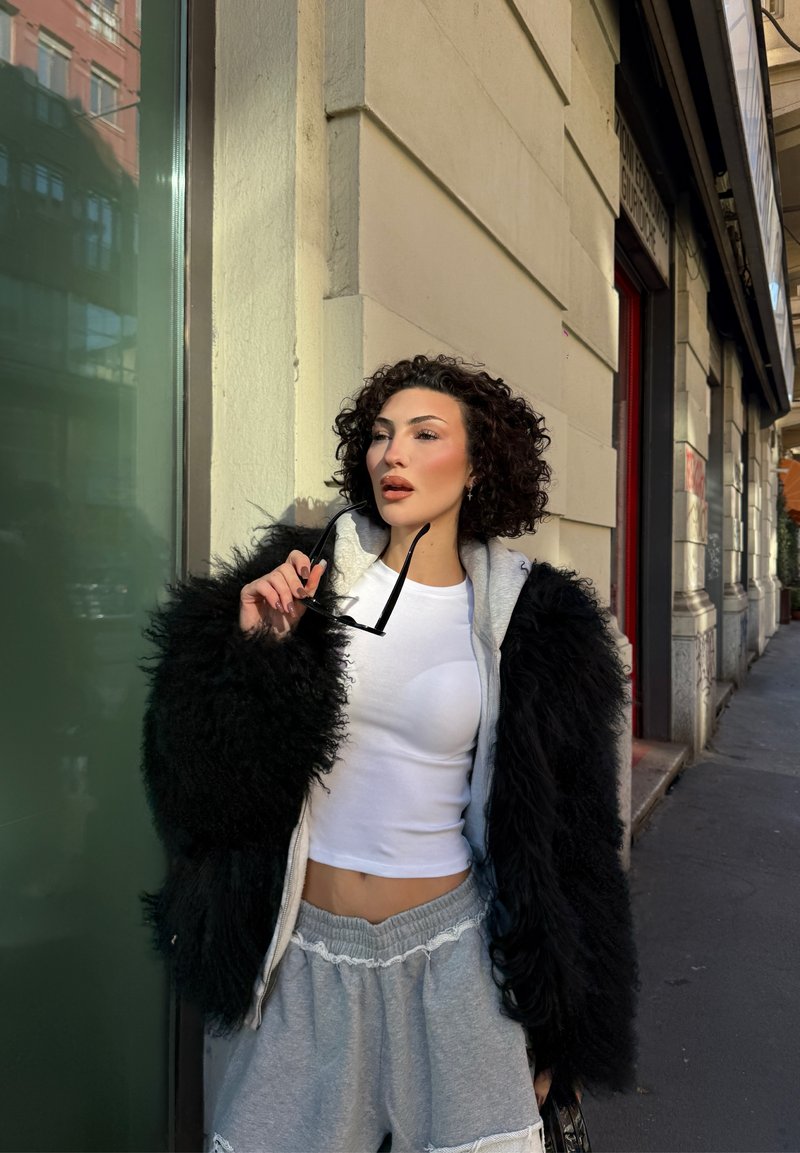 Schwarze Kunstpelzjacke mit grauer Kapuzenjacke darunter, weißes bauchfreies Top, graue Shorts, hält schwarze Sonnenbrille, steht draußen an einer Glaswand.