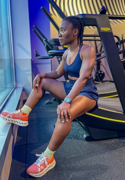 Atleta con un top deportivo azul oscuro y pantalones cortos, que lleva zapatillas de deporte coloridas con bandas. Sentada en el suelo del gimnasio junto a equipos de fitness.