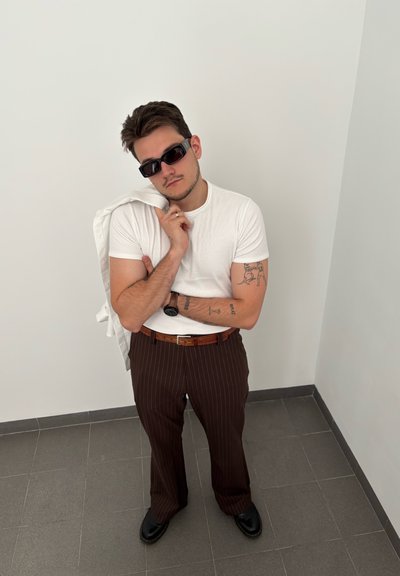 Camiseta blanca, pantalones marrones a rayas, zapatos negros y gafas de sol; sosteniendo una chaqueta blanca; tatuajes en los brazos visibles; entorno minimalista.