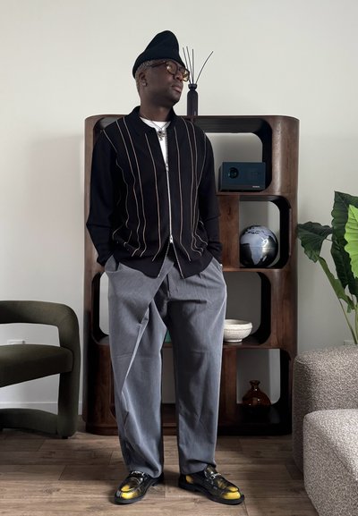 Hombre con gorro negro, gafas, suéter negro con cremallera y rayas verticales, pantalones grises y mocasines con detalles amarillos, de pie en el interior.