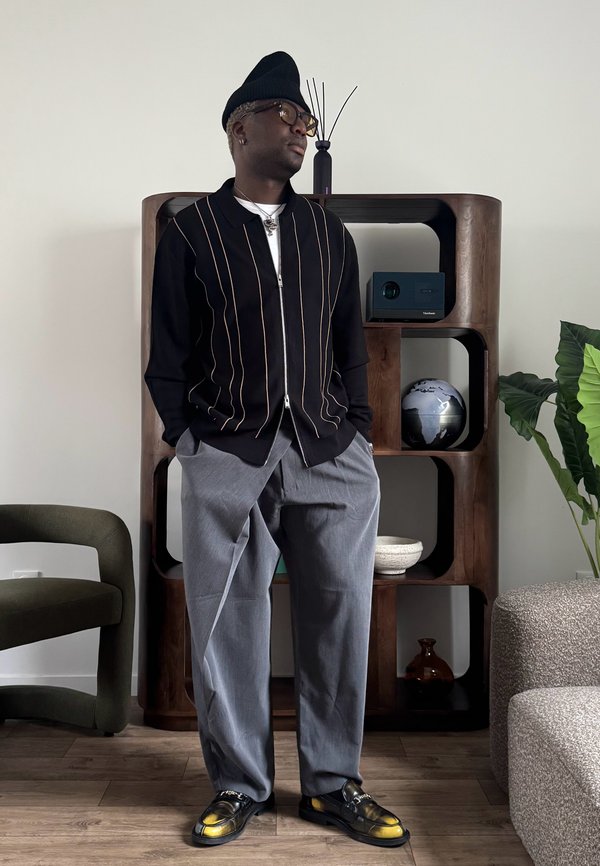 Moški, ki nosi črno kapa, očala, črni pulover na zadrgo z navpičnimi črtami, sive hlače in rumeno poudarjene loaferje, stoji v notranjosti.