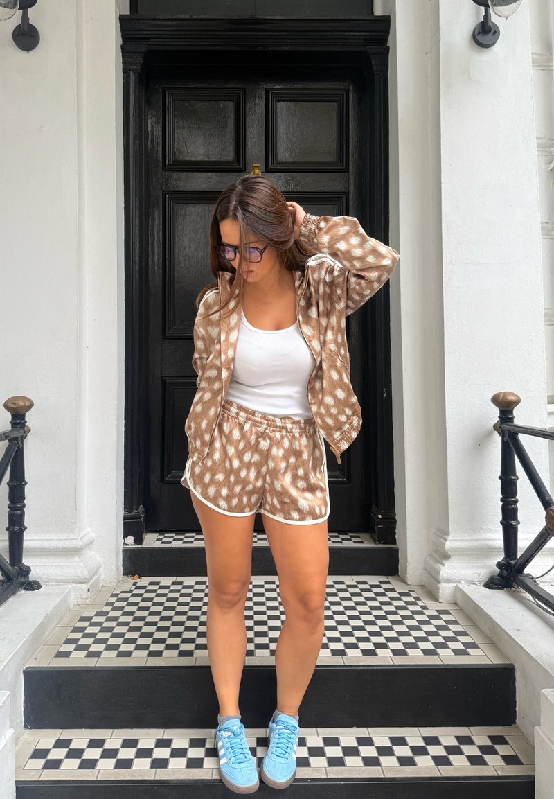 Young woman with brown spotted jacket and shorts, white tank top, blue sneakers stands on black and white tiled steps in front of black door.