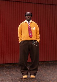 Homme portant un pull orange, une cravate rouge à motifs, un pantalon large marron, des bottes beige et un chapeau marron, debout contre un mur à panneaux rouges.