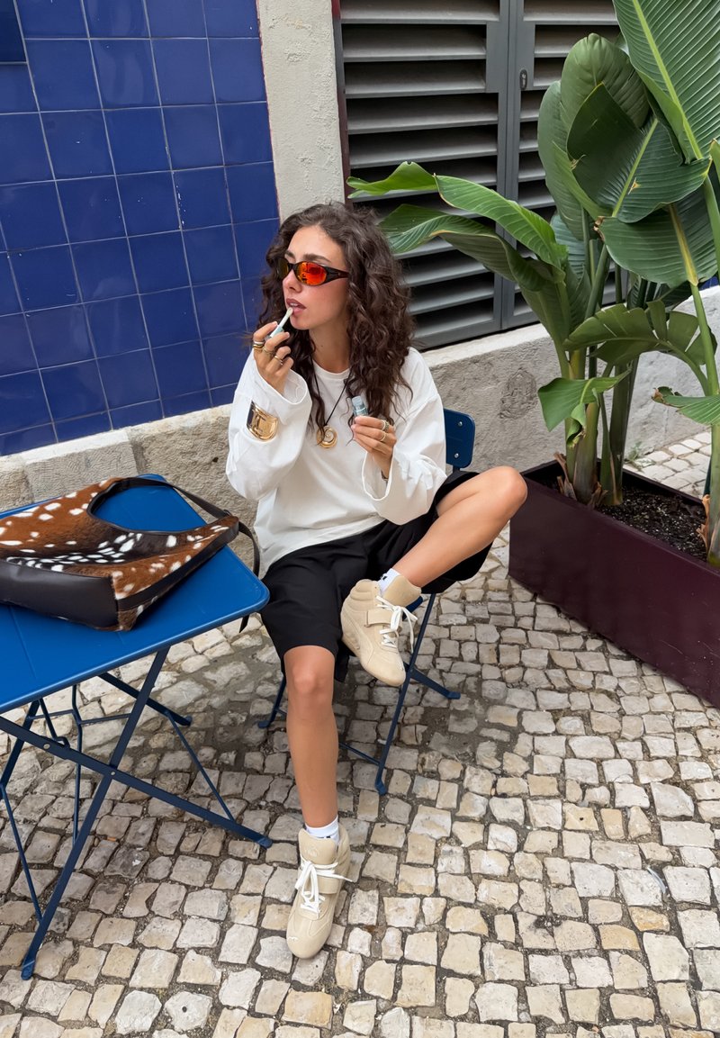 Woman with curly hair and sunglasses applies lip balm while sitting cross-legged on blue metal chair by table and plants outdoors.