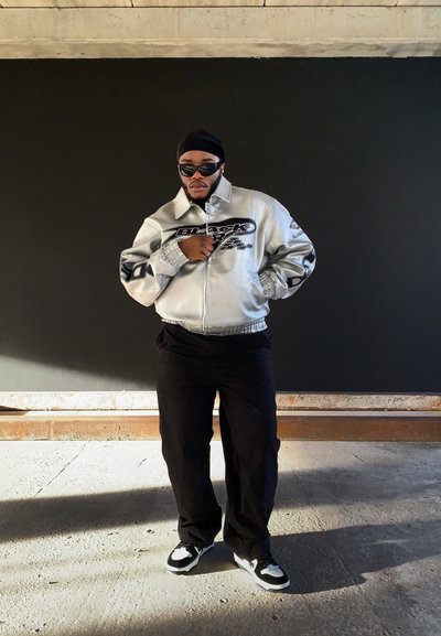 Chaqueta bomber plateada con gráficos negros, pantalones negros holgados y zapatillas en blanco y negro. Los accesorios incluyen gafas de sol y una cinta para la cabeza.