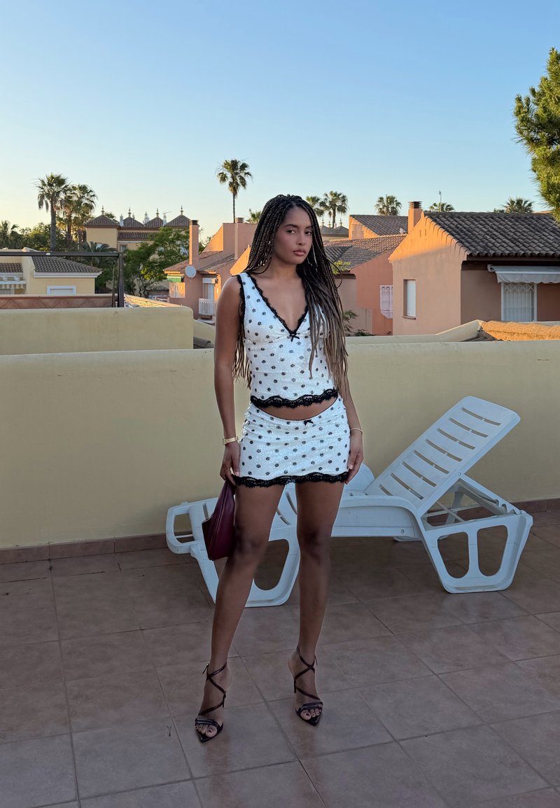 Femme aux longues tresses vêtue d'une robe blanche avec bordure en dentelle noire, debout sur une terrasse carrelée près de chaises longues blanches au coucher du soleil.