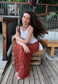 Žena sjedi na drvenoj klupi, odjevena u bijeli top bez rukava i dugu, crvenu, teksturiranu čipkastu suknju s valovitim uzorkom. Prirodno okruženje.