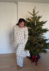 Bézs kockás pizsamaszett fekete fa mintákkal, laza szabású és gombos felsővel. Puha anyagból, bézs mamuszokkal viselve, egy fa mellett állva.