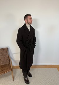 Homme portant un pardessus noir ceinturé, un col roulé beige, un pantalon noir et des chaussures habillées noires brillantes, debout à l'intérieur près d'une chaise en bois tressé.