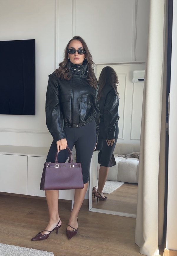 Woman in black leather jacket, black leggings, and sunglasses holds maroon handbag standing near full-length mirror in bright room.