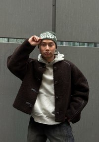 Ruda veltinio striukė su sagomis, dėvima ant šviesiai pilkos gaubtuotos bliuzono. Modelis dėvi žalią raštuotą kepuraitę, fone matomas miesto peizažas.