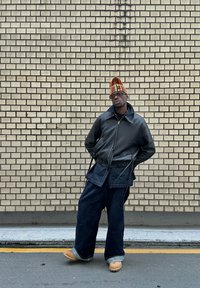 Svart läderjacka, oversized denimbyxor, bruna trätofflor och en färgglad randig hatt. Står mot en ljus tegelvägg.